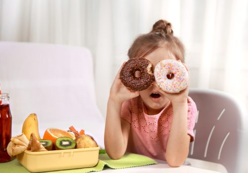 Little beautiful cheerful girl eating a donut and lunch box with fruits at home at the table, the concept of children's food